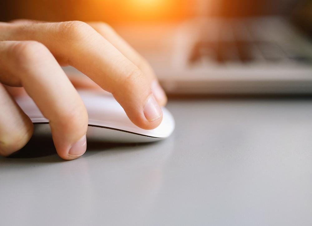 A close-up of a hand using a white computer mouse on a gray surface, with a blurred laptop in the background and a warm sunlight glow in the top corner.
