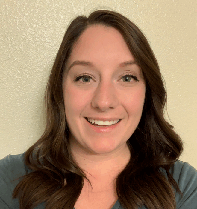 A woman with long brown hair and green eyes is smiling at the camera, standing against a plain, light-colored wall and wearing a teal top.