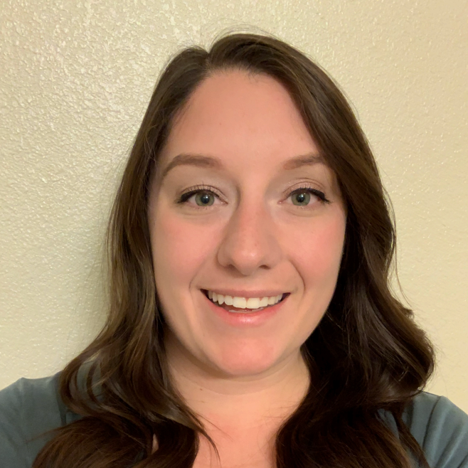 A woman with long brown hair and green eyes is smiling at the camera, standing against a plain, light-colored wall and wearing a teal top.