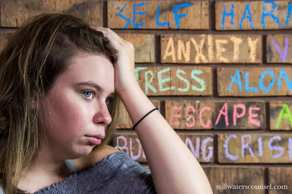 A woman looks pensive, resting her head in her hand. Behind her, a brick wall displays words like self harm, anxiety, and stress in colorful chalk. The image conveys themes of mental health struggles.