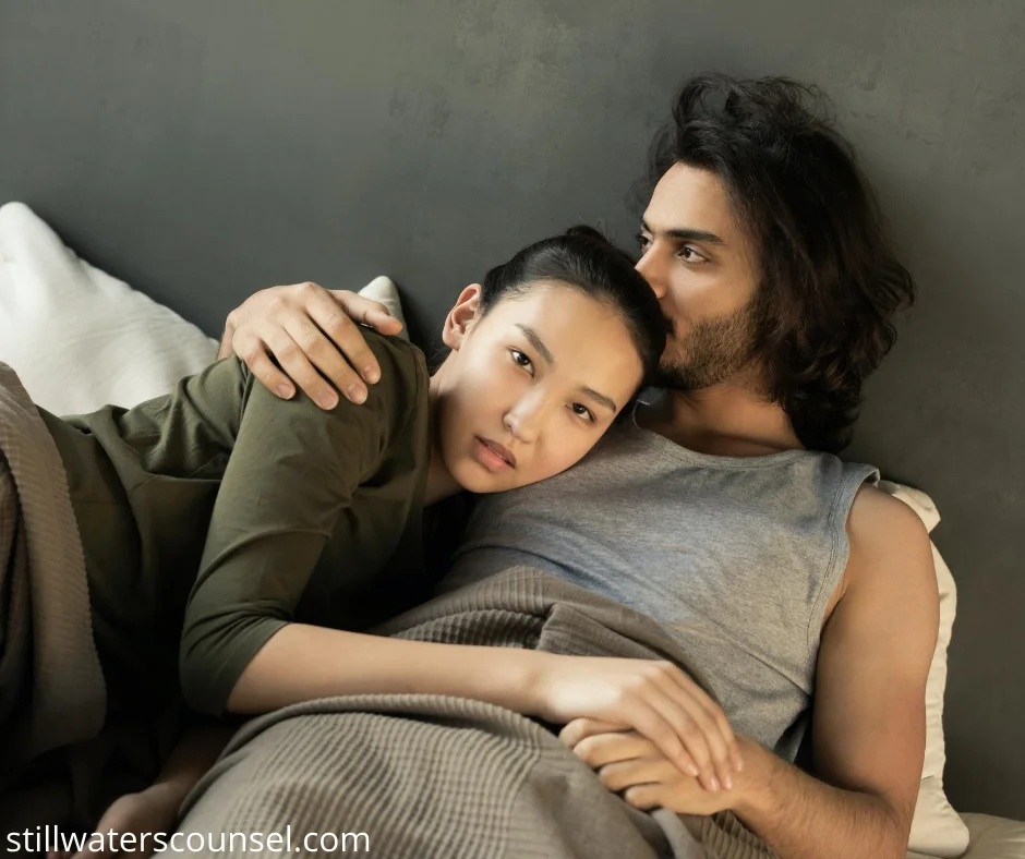 A couple lies on a bed, wrapped in a beige blanket. The woman rests her head on the mans chest, while he embraces her with a calm expression. Both are casually dressed, and the setting exudes warmth and intimacy.