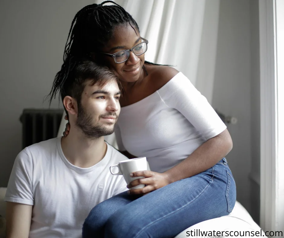 A man and woman sit together by a window, both wearing casual white tops. The woman is holding a mug and leaning affectionately on the mans shoulder, looking out. The atmosphere is calm and intimate.