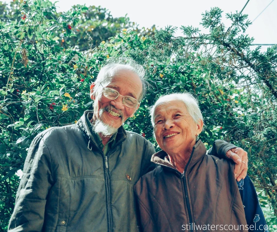 An elderly couple, smiling and embracing, stands in front of lush green foliage. The man wears glasses and a jacket, and the woman is dressed warmly. Sunlight filters through the leaves, creating a cheerful, serene atmosphere.