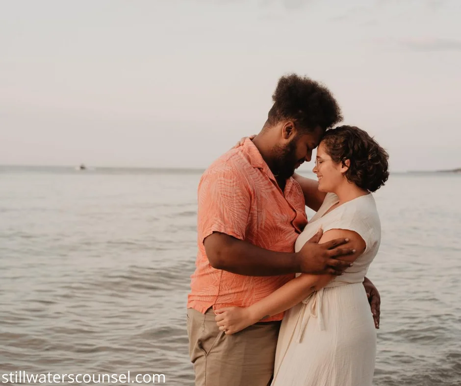 A couple embraces on a beach at sunset, standing close to the water with gentle waves in the background. The sky is light pink, and they appear peaceful and content.