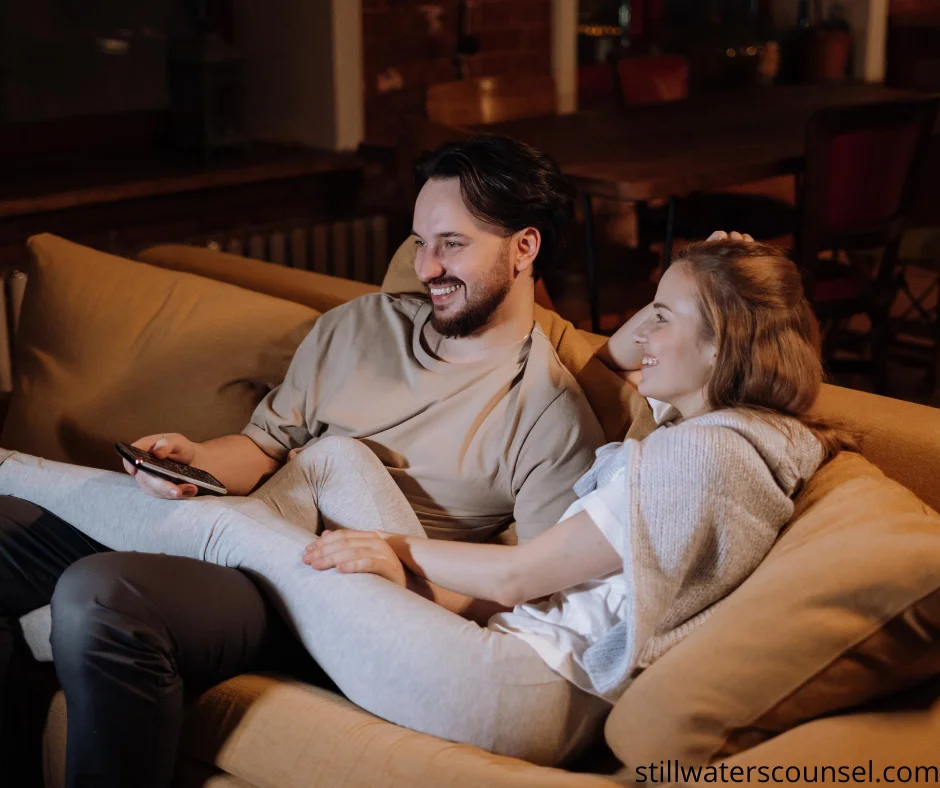 A smiling couple sits on a cozy couch, watching TV together. The man holds a remote control, and the woman rests her head on his shoulder, appearing relaxed and comfortable.