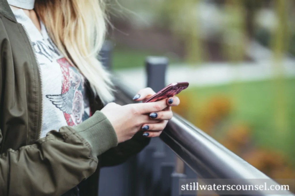 A person with long hair stands outdoors, using a smartphone. There wearing a casual outfit with a graphic t-shirt and a green jacket. The background is blurred, showing greenery and a railing.