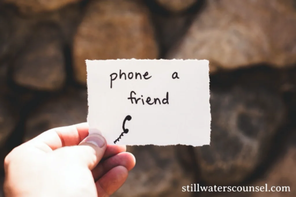 A hand holds a torn piece of paper with the handwritten phrase phone a friend and a drawing of a phone cord. The background features blurry rocks. The website URL stillwaterscounsel.com is in the bottom right corner.