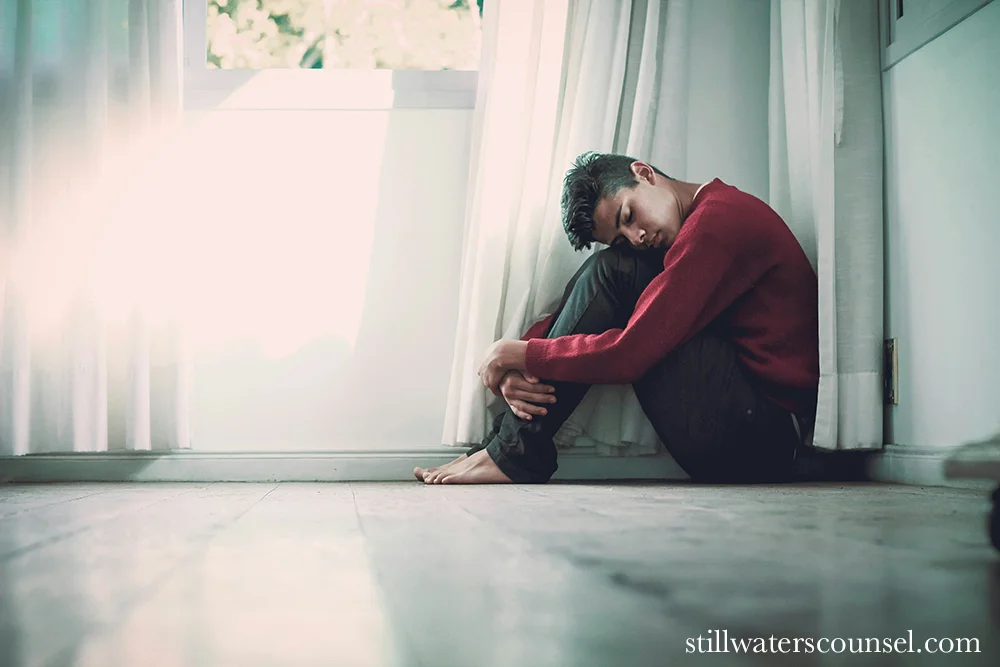 A person in a red sweater sits on the floor with knees pulled to their chest, leaning against a window with white curtains. The room is softly lit with sunlight filtering through. The expression is contemplative and introspective.