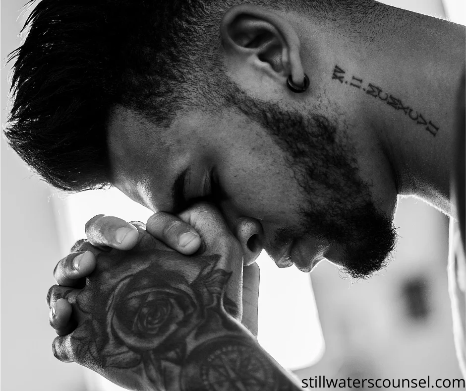 Black and white image of a man with a beard and tattooed forearm clasping his hands together in contemplation. A tattoo is visible on his neck. The atmosphere is introspective and peaceful.