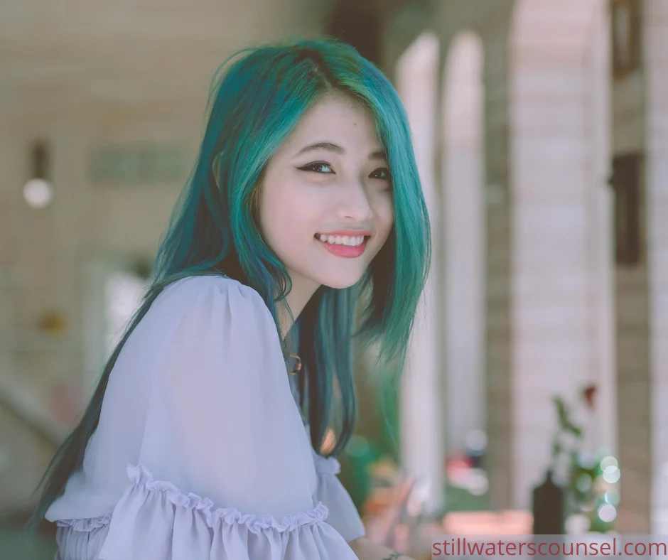 A woman with long turquoise hair smiles brightly while leaning on a surface. She is wearing a white, off-the-shoulder blouse, and the background is softly blurred, suggesting an indoor setting with archways.