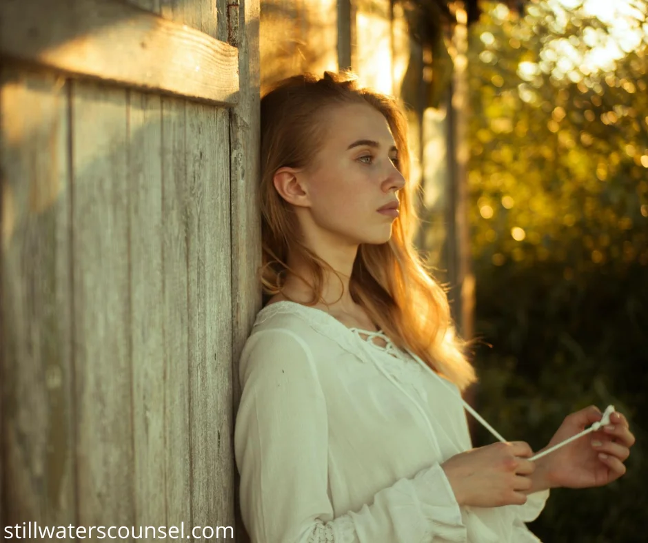 A woman with long hair stands against a wooden door, looking contemplatively into the distance. She is wearing a white blouse, with sunlight filtering through the trees in the background.