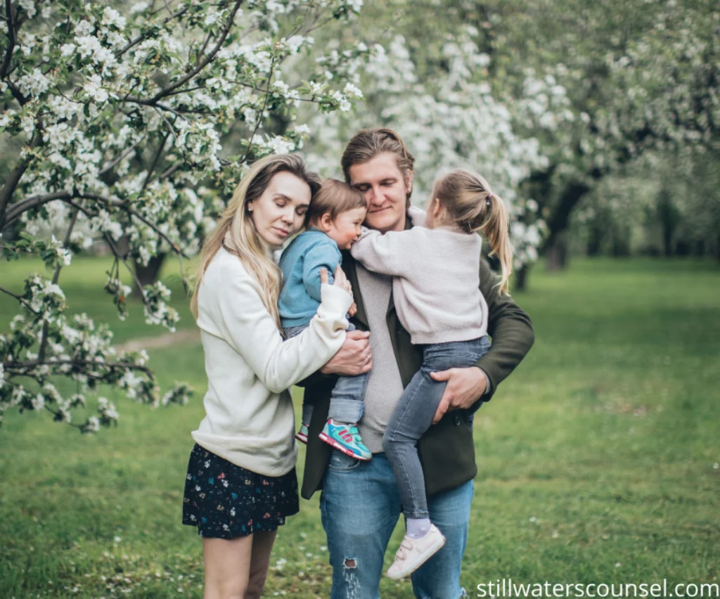 A family of four, standing together in an orchard with blooming trees. The parents are holding their children, who are dressed casually. The background features lush green grass and white blossoms on the trees.