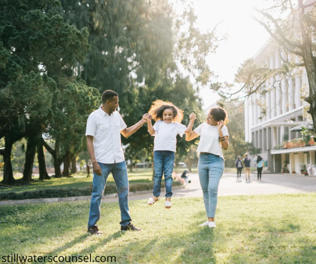 A family of three enjoying a sunny day at a park. The mother and father are holding hands with their child, who is joyfully jumping between them. The background features trees and a building.