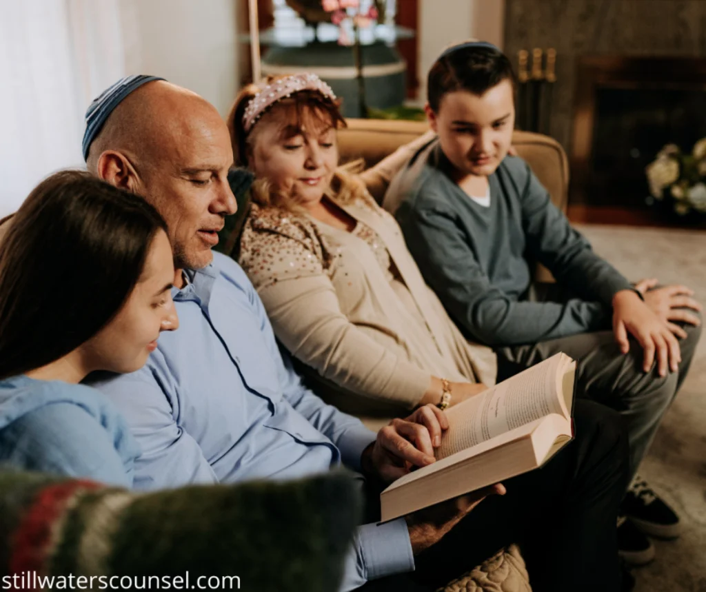 A family of four sits closely on a couch, engaged in reading a book together. The group includes two adults and two children, all wearing casual clothing. They appear to be in a cozy living room setting.