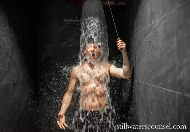 A person stands under a waterfall shower, water cascading over their head and shoulders, in a dark tiled room. Their mouth is open, and they hold a rope attached to the shower. The image has the website watermark stillwaterscounsel.com.