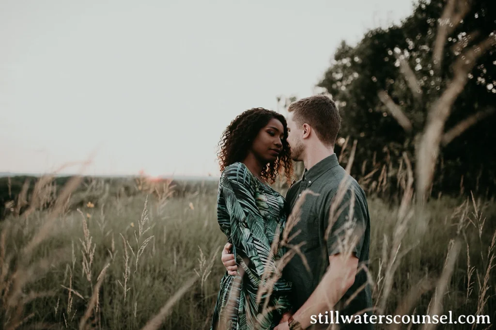 A couple standing closely in a field of tall grass at sunset, embracing gently while looking into each others eyes. The sky is dimly lit, creating a serene and intimate atmosphere.