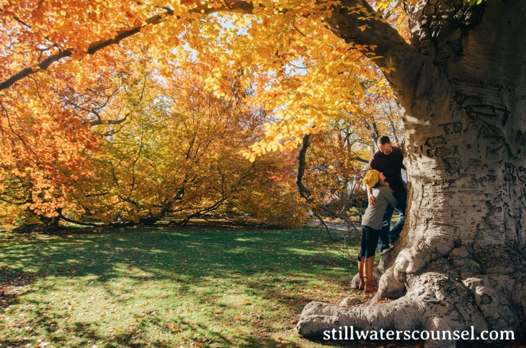 A couple leans against a large tree with carved initials, surrounded by vibrant autumn foliage in a sunlit park. The ground is covered with fallen leaves, creating a warm, colorful scene.