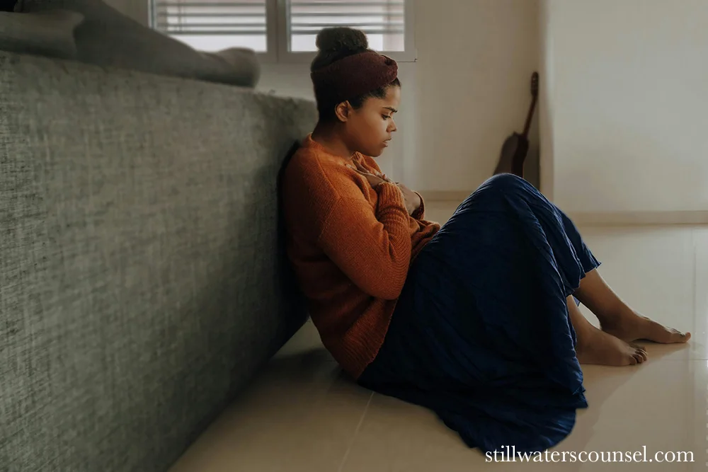 A person sitting on the floor, leaning against a couch, wearing a brown sweater and blue skirt. They appear deep in thought or contemplation. The room has soft natural lighting and a guitar leaning against the wall in the background.