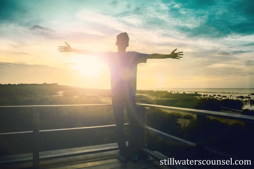 A person stands on a deck with arms outstretched, facing a sunrise or sunset over a scenic landscape of greenery and water. The sky is filled with clouds and golden light. Text at the bottom reads stillwaterscounsel.com.