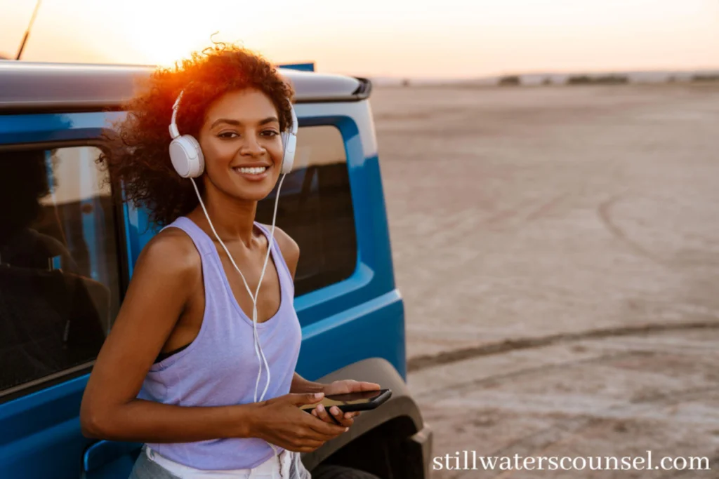 A woman with headphones leans against a blue vehicle in a desert landscape at sunset, holding a smartphone and smiling. The website stillwaterscounsel.com is visible.