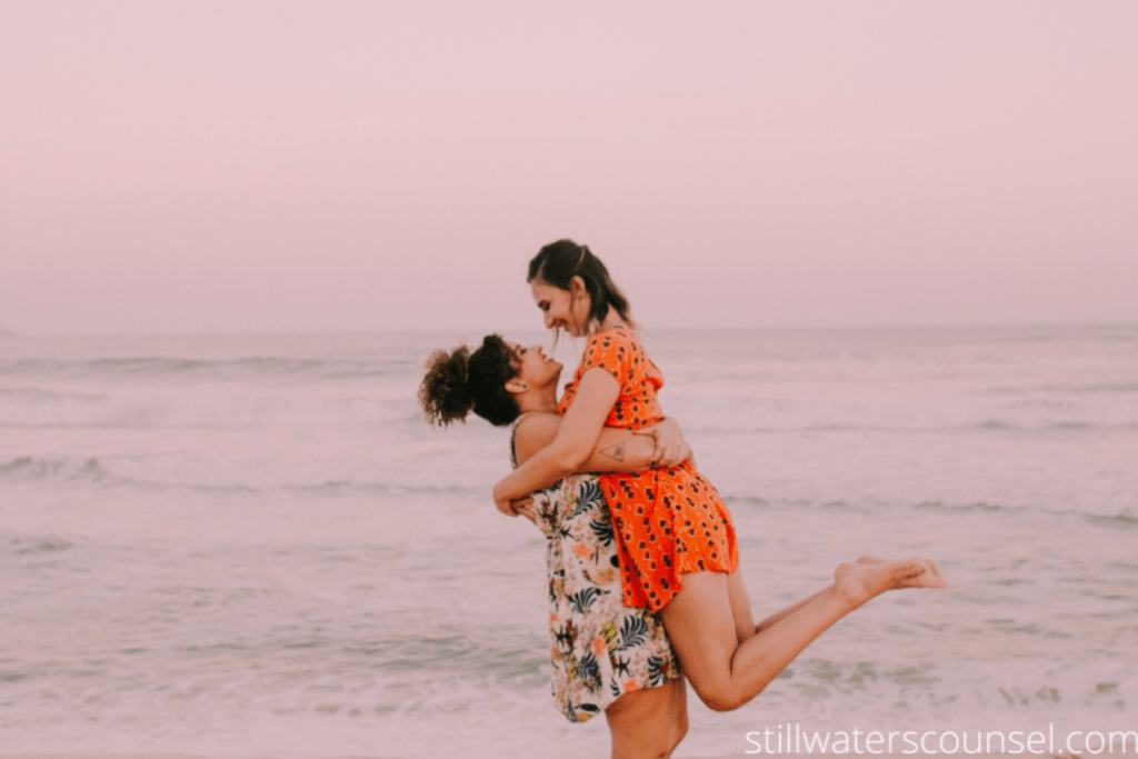 Two people joyfully embrace at the beach during sunset. One person lifts the other in the air. They are both wearing colorful dresses, with waves gently rolling in the background. The sky is a soft pink hue.
