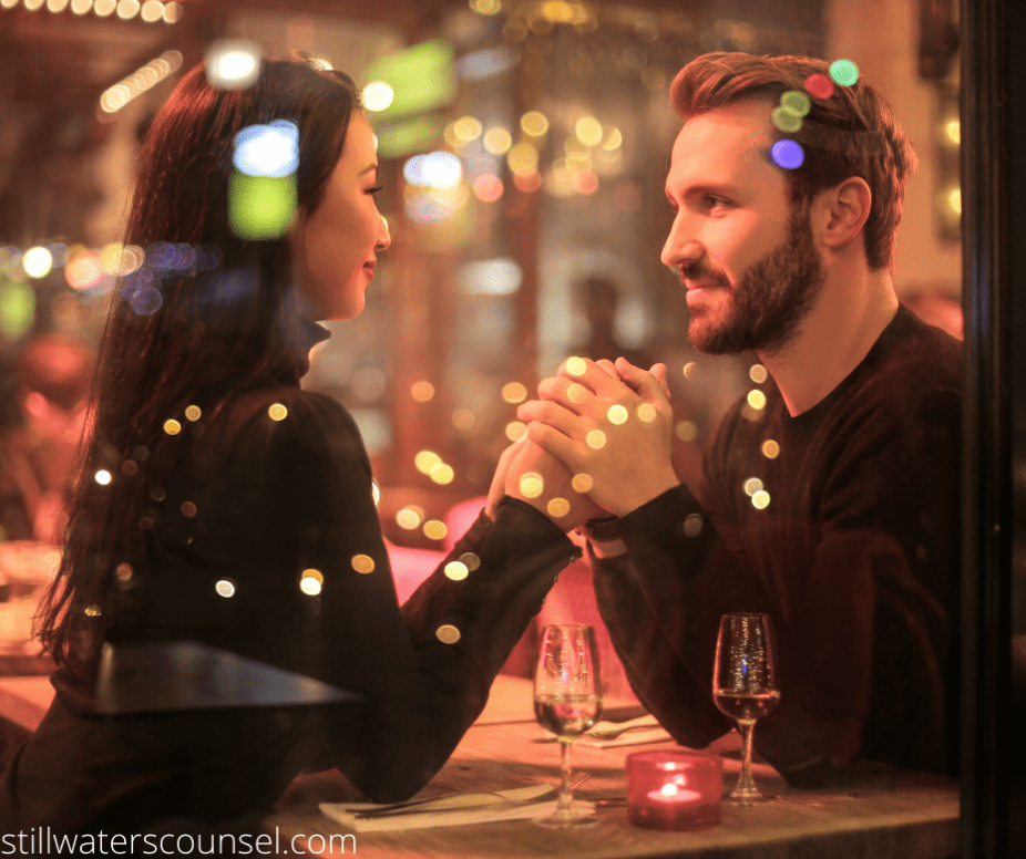 A couple sits at a table holding hands in a warmly lit restaurant, with glasses of wine in front of them. They gaze at each other through a window, surrounded by soft, glowing lights, creating a romantic atmosphere.