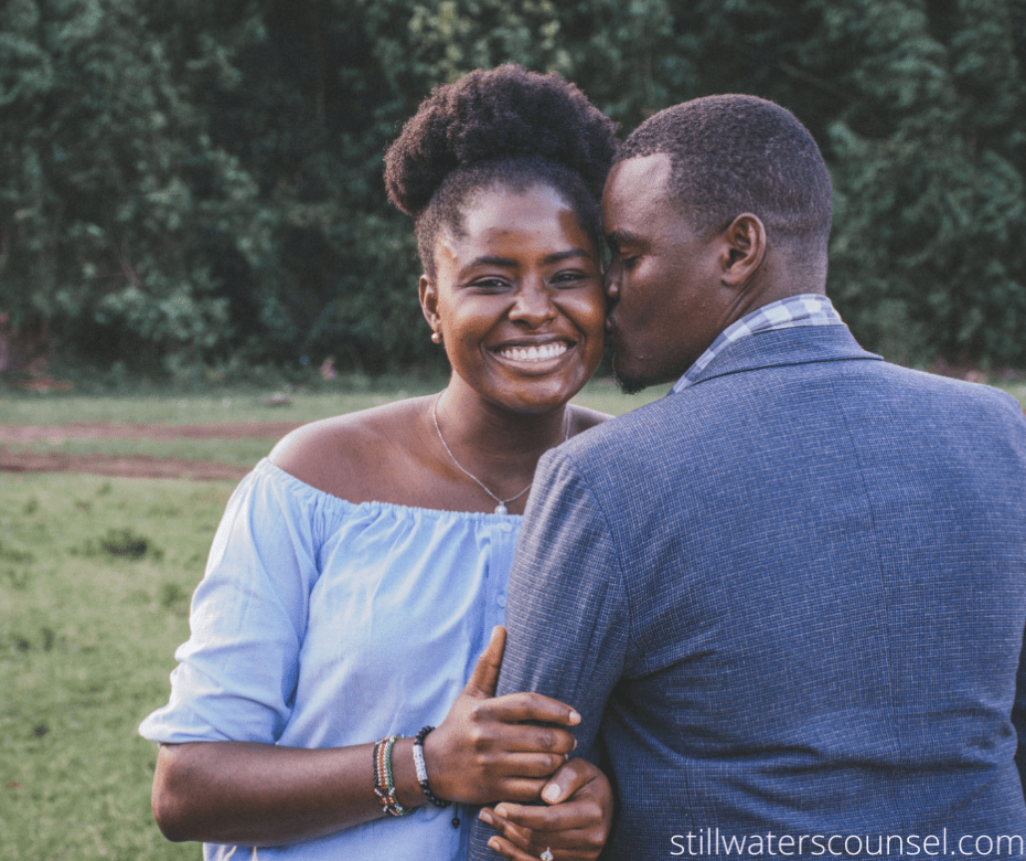 A man and woman stand outdoors, with the man kissing the womans cheek. The woman is smiling broadly, wearing an off-the-shoulder blue blouse, and the man is in a blue jacket. They are in a green field with trees in the background.