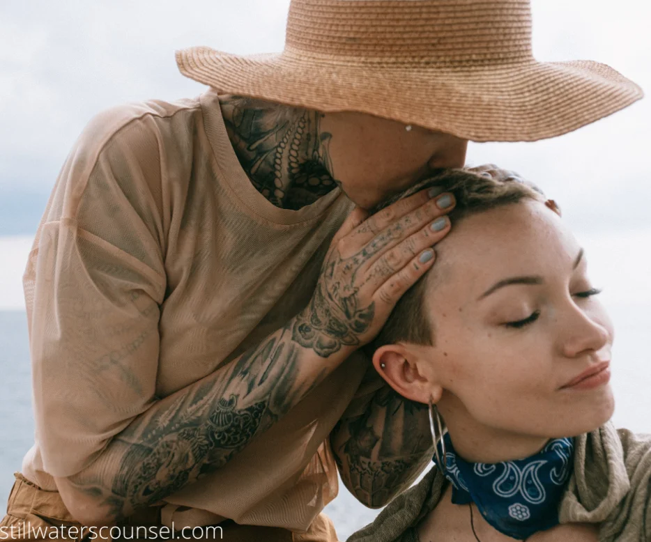 A person in a wide-brimmed hat kisses the head of another person with short hair, who is wearing a blue bandana. Both have visible tattoos and piercings. The background is a soft, blurred outdoor setting.
