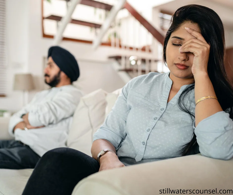 A woman sits on a couch with her hand on her forehead, appearing stressed, while a man with a turban sits in the background with arms folded, looking away. The room has a staircase and soft lighting.