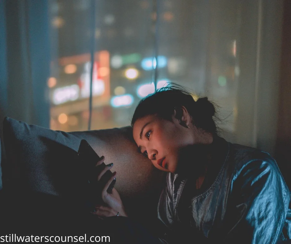 A woman lies on a couch, looking at her smartphone. The room is dimly lit, with city lights visible through the window in the background, creating a calm and introspective atmosphere.