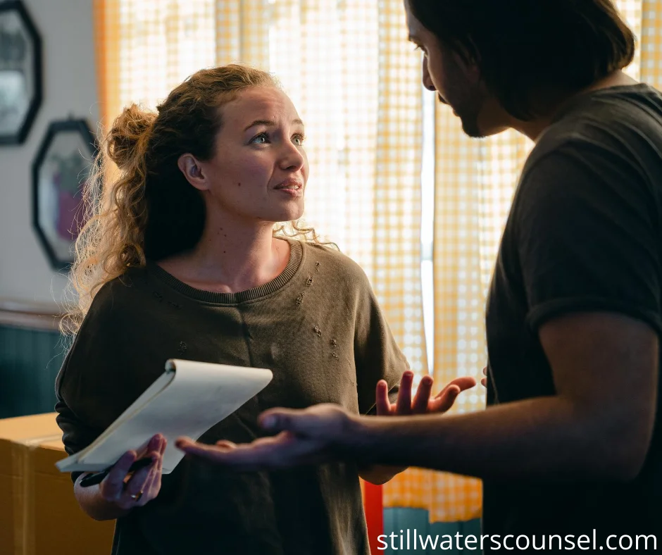 A woman with curly hair, holding a notepad, looks concerned while talking to a man with long hair. They are indoors with checkered curtains in the background. The man gestures with his right hand.