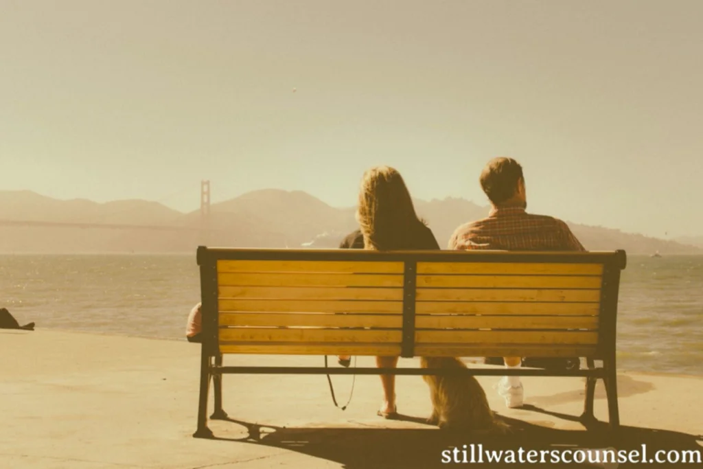 A couple sits on a wooden bench, facing a body of water with a distant view of a bridge and mountains. The scene is serene and sepia-toned, suggesting a nostalgic or relaxed atmosphere. The bench is on a concrete surface near the waters edge.