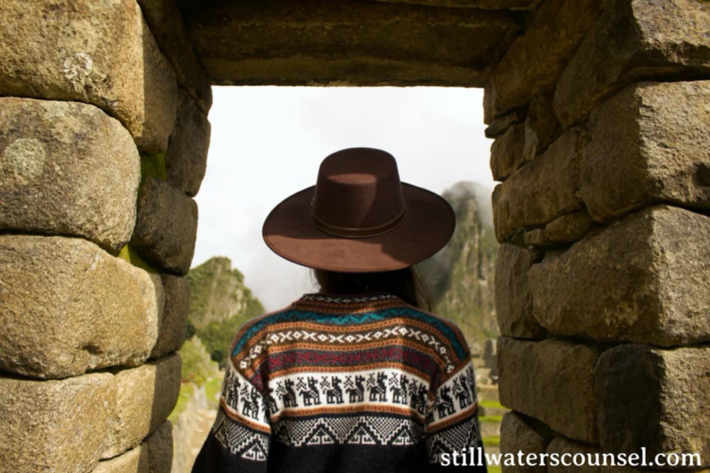 A person wearing a wide-brimmed brown hat and a colorful, patterned sweater stands at a stone archway, looking out at an ancient ruin. The persons back is to the camera. Rock formations are visible through the arch. URL in the corner says stillwaterscounsel.com.