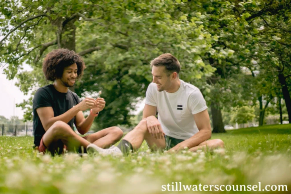 Two people sit on grass in a park, engaged in conversation and smiling. They appear relaxed, surrounded by lush greenery on a sunny day. The text stillwaterscounsel.com is visible at the bottom.