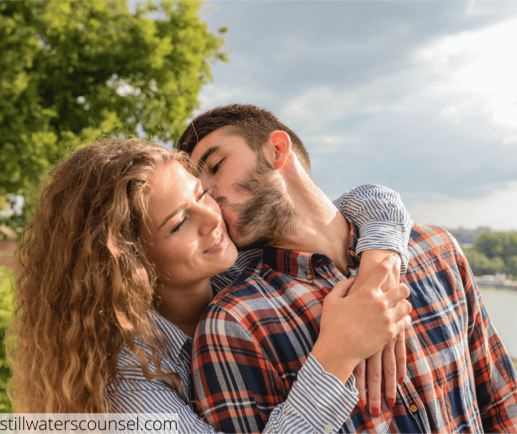 A couple stands outdoors on a sunny day, with trees and a river in the background. The woman embraces the man from behind while he kisses her cheek. They both wear casual plaid shirts and appear happy.