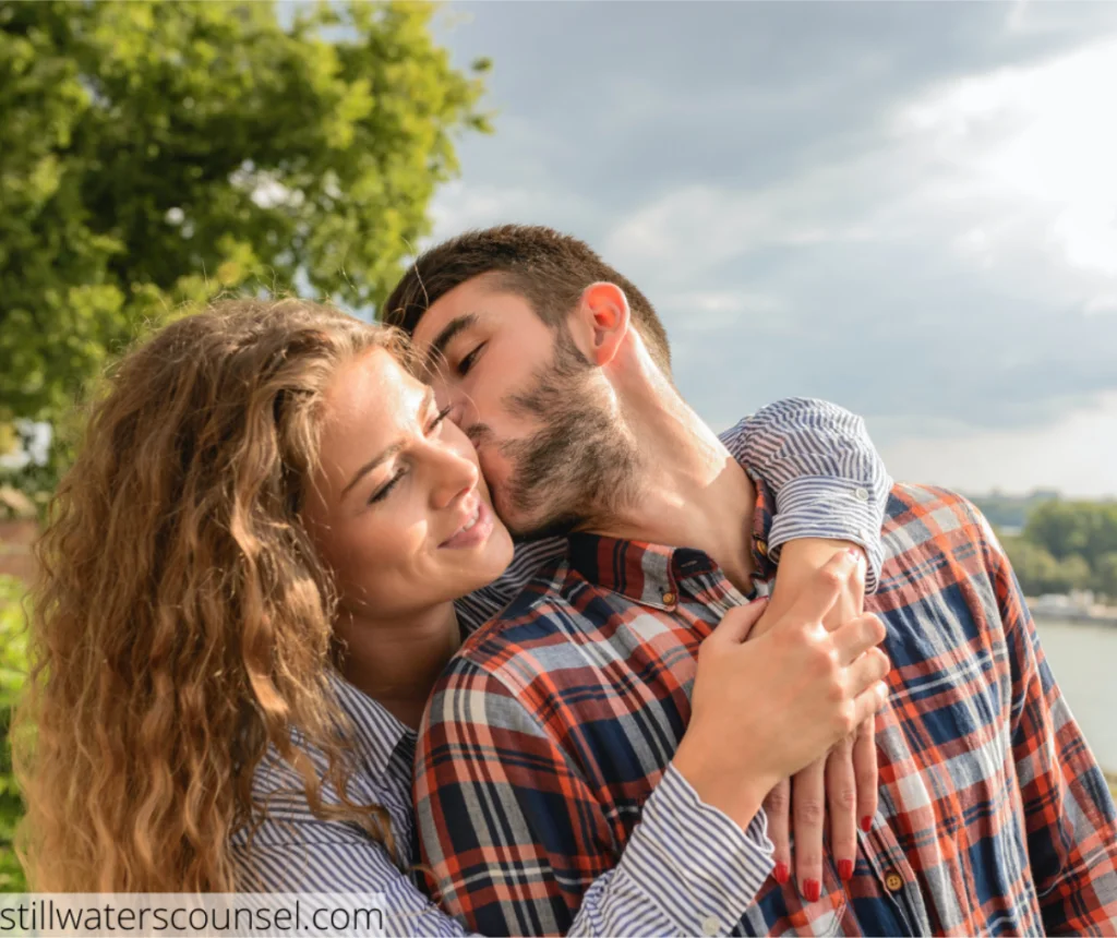 A couple stands outdoors on a sunny day, with trees and a river in the background. The woman embraces the man from behind while he kisses her cheek. They both wear casual plaid shirts and appear happy.