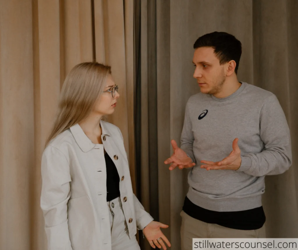 A woman with long blonde hair and glasses in a light-colored outfit and a man in a grey sweater are having a serious conversation, gesturing with their hands. They stand in front of beige and brown curtains.