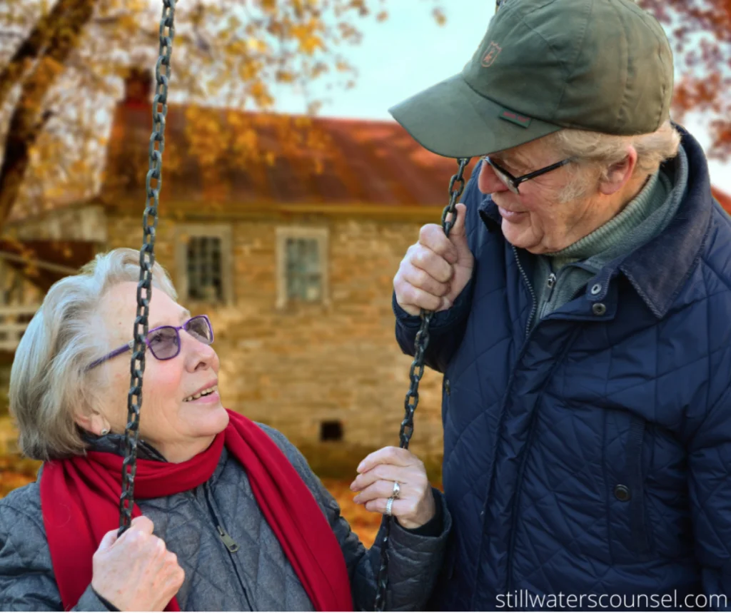 An elderly couple smiles at each other while standing next to a swing. They are dressed warmly with coats and scarves. A rustic building and autumn trees are in the background.