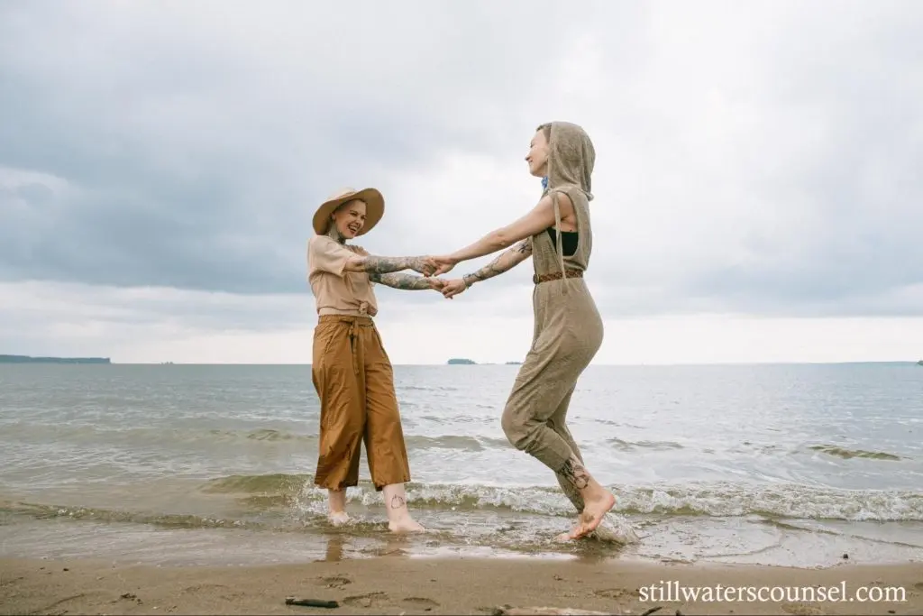 Two people joyfully hold hands and dance at the edge of the sea. The person on the left wears a wide-brimmed hat and brown pants, while the person on the right wears a sleeveless hooded outfit. The sky is cloudy, creating a serene atmosphere.