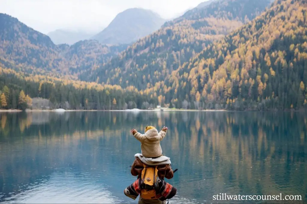 A person carrying a child stands by a serene lake with clear reflections of surrounding autumn trees and mountains. The child is wearing a yellow hat and has arms outstretched. The scene conveys a sense of freedom amidst nature.