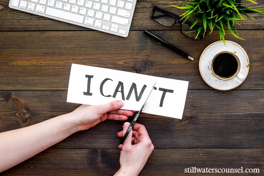 A person cuts a paper with I CANT written on it, removing the T to change it to I CAN. The scene includes a keyboard, glasses, a plant, and a cup of coffee on a wooden desk.