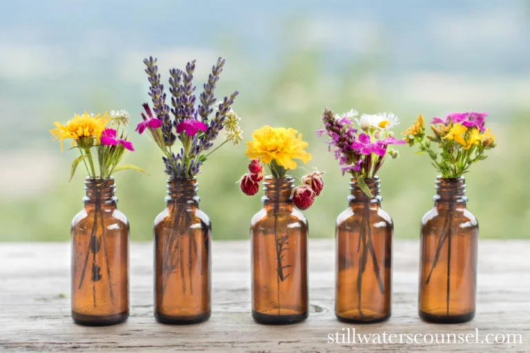 Five small brown glass bottles on a wooden surface, each holding a bouquet of colorful wildflowers, including lavender, daisies, and other blooms, against a blurred natural background.