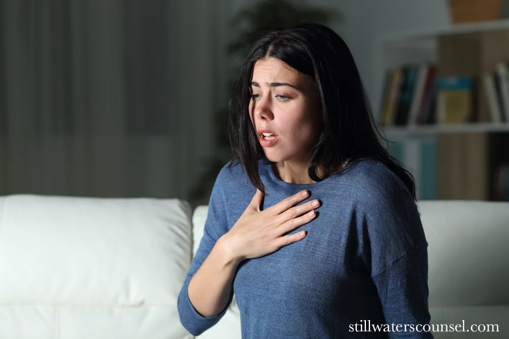 A woman with long dark hair, wearing a blue sweater, sits on a white couch appearing anxious and holding her chest with one hand. The room is dimly lit, with a bookshelf in the background.