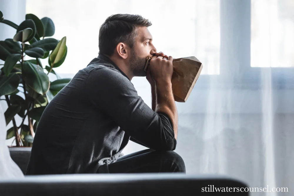 A man sits on a couch, using a brown paper bag for breathing. He appears deep in thought or anxious. A large leafy plant is visible nearby. Natural light filters through a window in the background, creating a serene atmosphere.