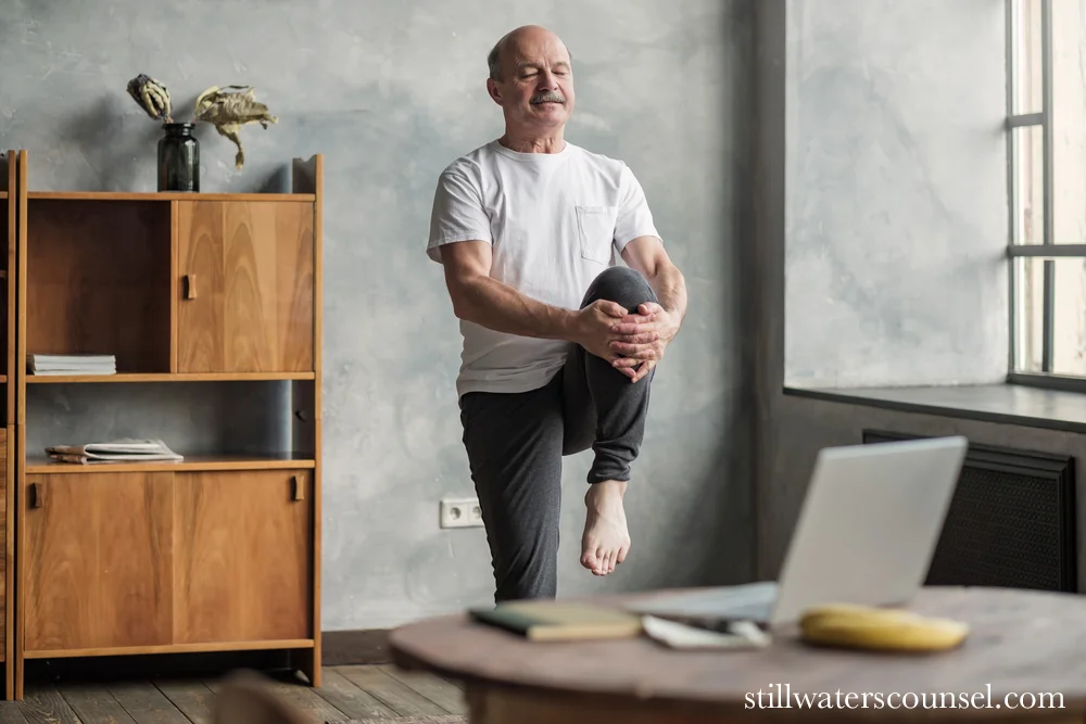 An elderly man in a white t-shirt and gray pants performs a balance exercise by holding one knee up with both hands. He stands in a room with a wooden bookshelf and a table with a laptop. Natural light filters through a window.