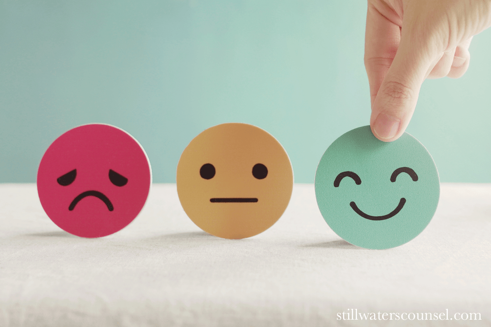 A hand selects a green smiley face disc from a row of three emoji discs, with a sad red face and a neutral orange face also visible against a soft blue background.