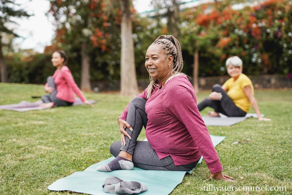 A group of women practicing yoga outdoors on a grassy area. The woman in the foreground is wearing a pink top and gray leggings, sitting in a yoga pose with one leg crossed. Trees and flowers are in the background.