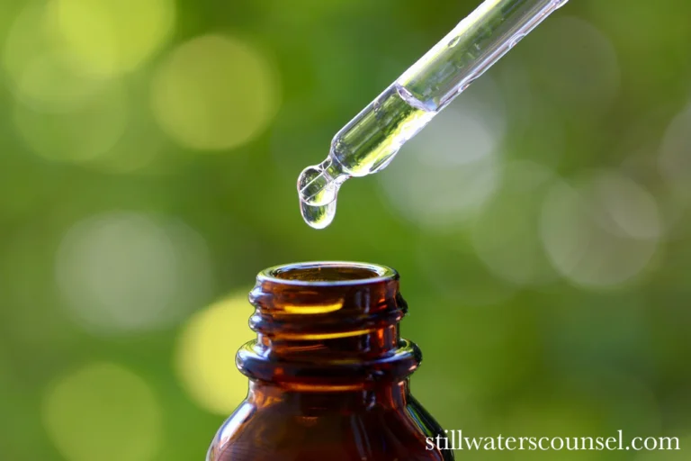 A close-up of a dropper releasing a drop of liquid into a small brown glass bottle. The background is blurred with greenery and bokeh effects, creating a serene, natural atmosphere.