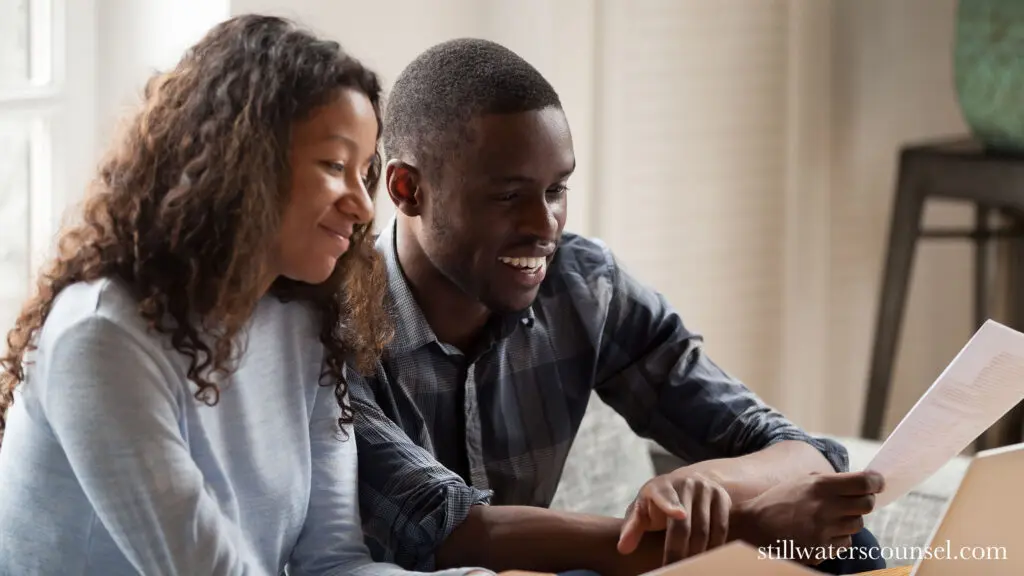 A smiling couple sits together on a couch, looking at a document. They both appear engaged and content. Light filters through the window behind them, creating a warm and inviting atmosphere.