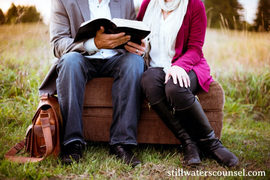 A couple sits on a couch in a field, with a man holding an open book and a woman sitting close. Both are dressed casually, with a brown bag on the ground beside them. The website stillwaterscounsel.com is visible in the corner.